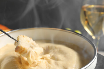 Dipping piece of bread into fondue pot with melted cheese on grey background, closeup
