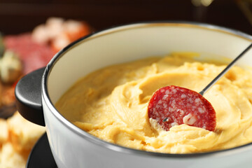 Dipping piece of sausage into fondue pot with melted cheese on blurred background, closeup