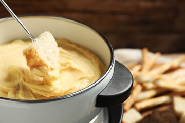 Dipping piece of bread into fondue pot with melted cheese on blurred background, closeup