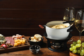 Fork with piece of sausage, melted cheese in fondue pot and other products on wooden table. Space...