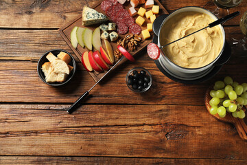 Fork with piece of sausage, melted cheese in fondue pot and other products on wooden table, flat...