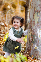 Smiling little girl standing by a tree, touching its bark, leaves at their feet