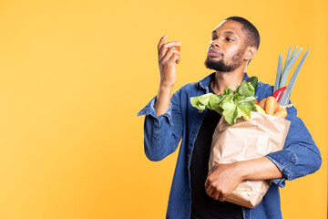 African american guy admiring a tomato and smelling the fresh aroma against yellow background,...