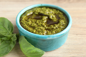 Tasty pesto sauce in bowl and basil on wooden table, closeup