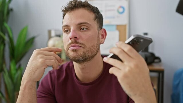 Young hispanic man with a beard having a phone conversation in a modern office setting, looking confused.