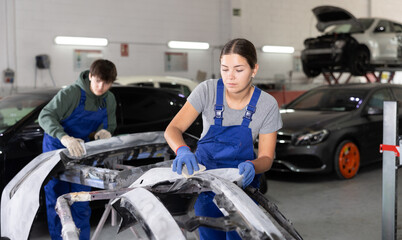 Young female auto mechanic in blue overalls preparing car for painting, sanding removed bumper with sandpaper after applying putty in specialized garage, focused on work