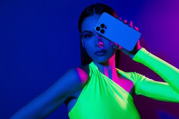 Portrait of gorgeous confident model girl hand hold smart phone cover one eye isolated on blue neon...