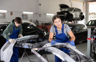 Young female auto mechanic in blue overalls preparing car for painting, sanding removed bumper with...