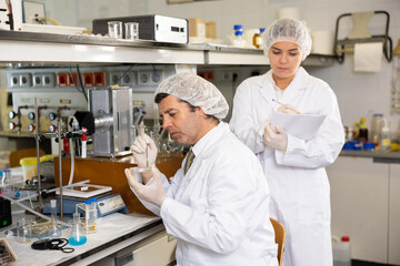Middle-aged male chemist examining solution in petri dish while female writing report in research...