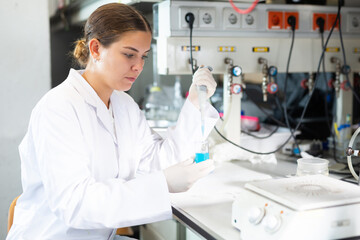 Focused female chemist pipetting blue reagent into solution in test tube while performing experiments in research center
