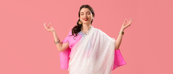 Beautiful young Indian woman in sari meditating on pink background