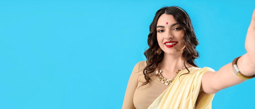Beautiful young Indian woman in sari taking selfie on blue background
