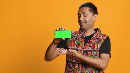 Content creator presenting green screen mobile phone, isolated over studio background. Indian...