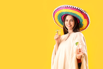 Beautiful young Mexican woman in sombrero and with tequila on yellow background