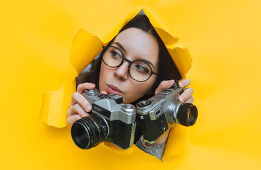 A paparazzi girl in glasses with two old SLR cameras looks out from hiding place and carefully observes what is happening. Yellow paper, torn hole. Tabloid press. Looking for a theme for stock photos.