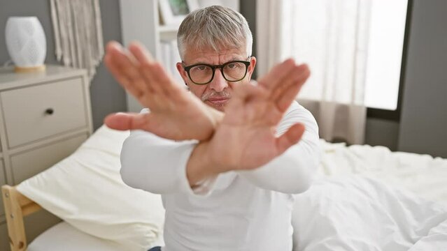 Grey-haired, middle-aged man in pyjamas angrily rejects, wearing a stern expression and crossed arms in the bedroom