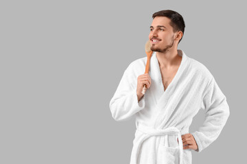 Handsome young man in bathrobe with massage brush on grey background