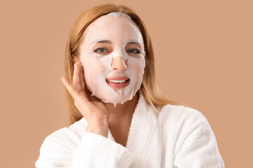 Mature woman in bathrobe with facial sheet mask on beige background, closeup