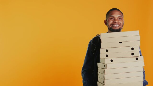 African american pizzeria deliveryman holding stack of pizza boxes to deliver food order to customer in studio. Friendly takeout service employee carries takeaway meal package. Camera B.
