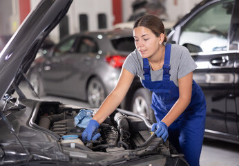 Young female mechanic in uniform repairs under hood of car in car service station