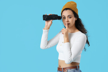 Young African-American woman with binoculars showing silence gesture on blue background