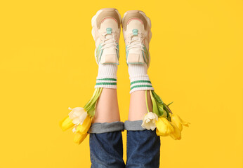 Legs of beautiful young woman in sneakers with tulips on yellow background