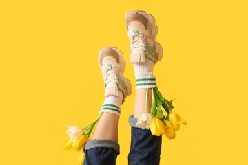 Legs of beautiful young woman in sneakers with tulips on yellow background