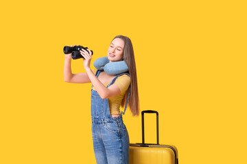 Young woman staying near her luggage and looking in binoculars on yellow background