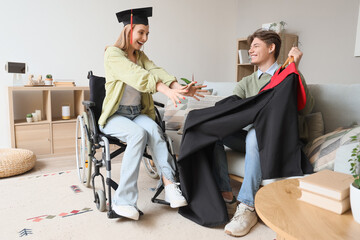 Female graduate in wheelchair and her friend with robe at home