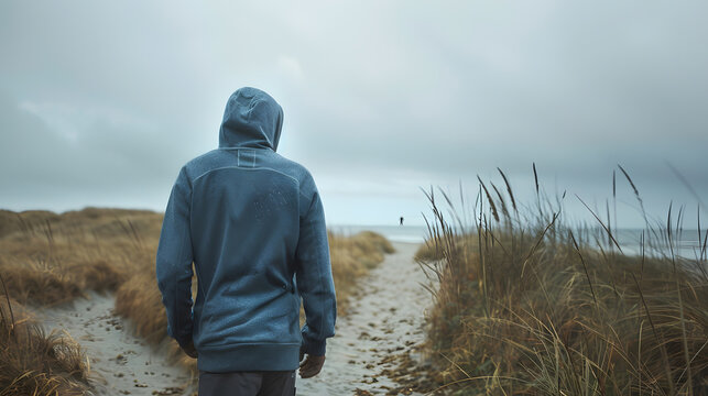 An adult male with a hoodie walking along a sandy path by the shore on a cloudy day : Generative AI