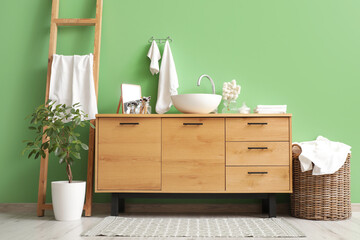 Wooden chest of drawers with sink  and laundry basket near green wall in modern bathroom