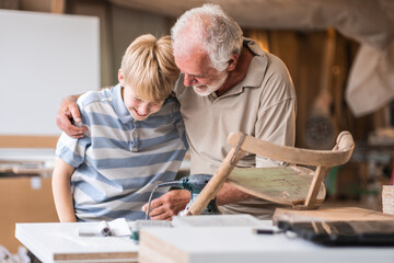 A granddad teaches his grandson how to fix furniture in his carpenter workshop, hugging him as they...