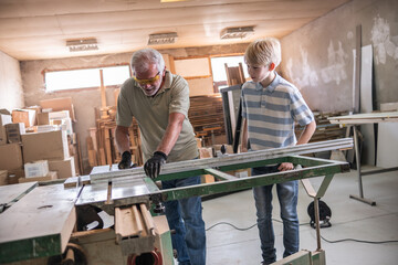 Granddad and grandson work together in the carpenter workshop, learning how to use an electric circular saw. Granddad guides his grandson through the process, teaching him the skills and safety measur