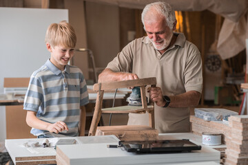 A grandfather teaches his grandson how to fix furniture in his carpenter workshop. They work...