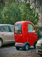 A red retro mobility scooter with a rounded roof is parked next to a silver car, set against a backdrop of trees and bushes, blending modern and vintage elements seamlessly