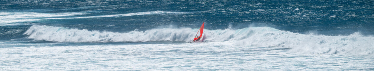 Obraz premium Man on a wind surfer with red sail crashing on a wave on the Pacific Ocean off the coast of Maui at Hookipa Beach, Hawaii 