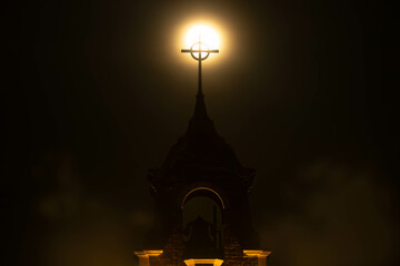 Full moon on a dark and gloomy night. The glowing planet rises behind a steeple silhouette of a Christian church. Lights glow on the bell tower of the place of worship