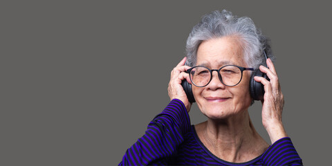 Cheerful senior woman wearing wireless headphones with a smile while standing on a gray background.