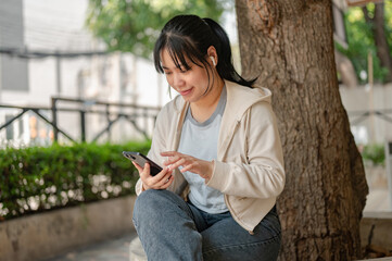 Young Asian woman wearing earbuds, using her phone, listening to music while chilling in a city park