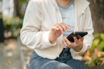 A cropped shot of a young Asian woman sits on a bench in a city park and uses her smartphone.