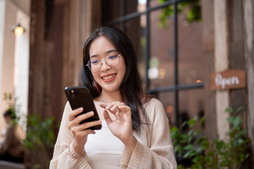 A young Asian woman sits at an outdoor table of a cafe and uses her phone to read something online.