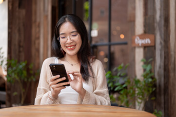 A young Asian woman sits at an outdoor table of a cafe and uses her phone to read something online.