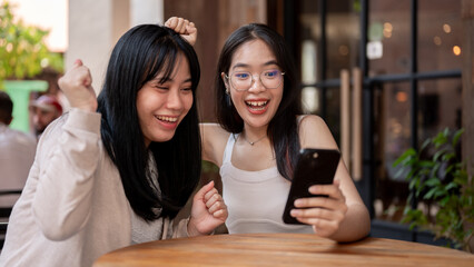 Two excited Asian female friends are looking at a smartphone, celebrating good news together.