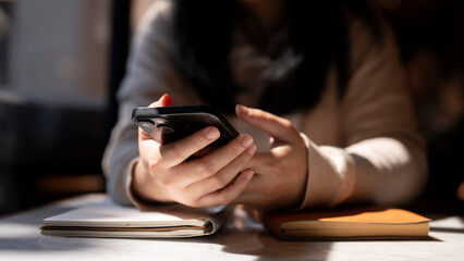 A cropped image of a woman using her smartphone at a table by the window on a sunny day.