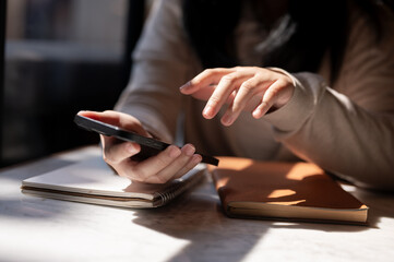 A cropped image of a woman using her smartphone at a table by the window on a sunny day.