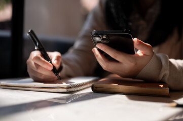 A woman holding a smartphone and taking notes or making list in a notebook at a table indoors.