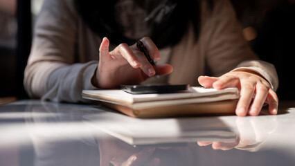 A cropped image of a woman holding a pen and using her smartphone at a table.
