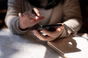 A cropped image of a woman holding a pen and using her smartphone at a table.