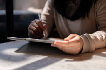 A cropped image of a woman holding a stylus pen, writing on a digital tablet, sitting at a table.