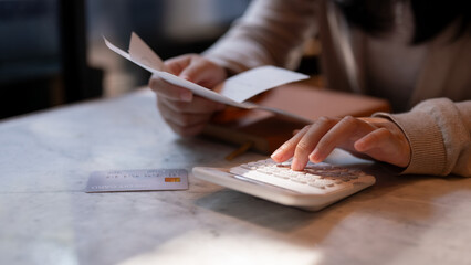 A woman holding receipt and using a calculator, sitting at a table indoors, calculating her expenses